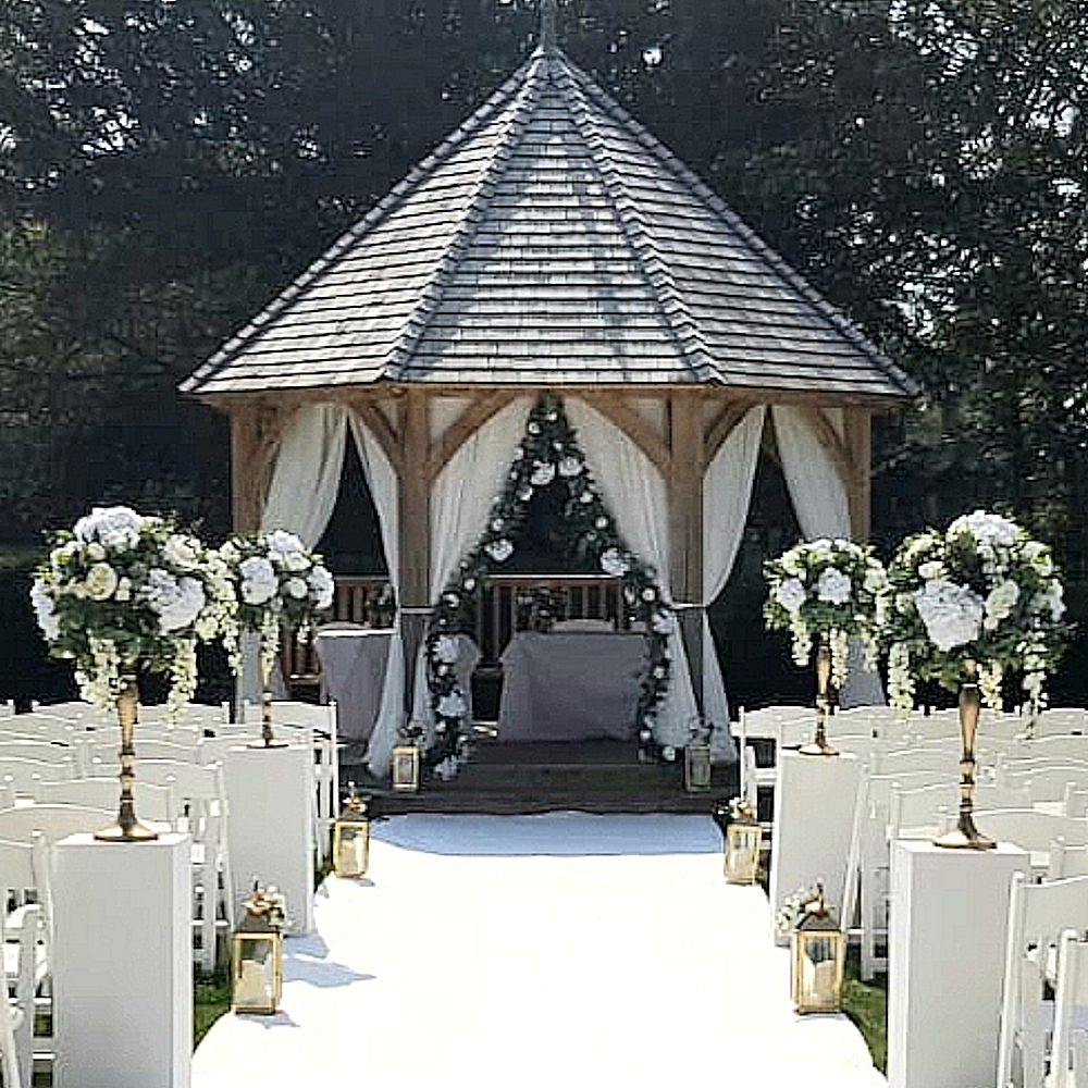 Gazebo white plinths chiffon drapes foliage garland white carpet white folding chairs down hall ceremony